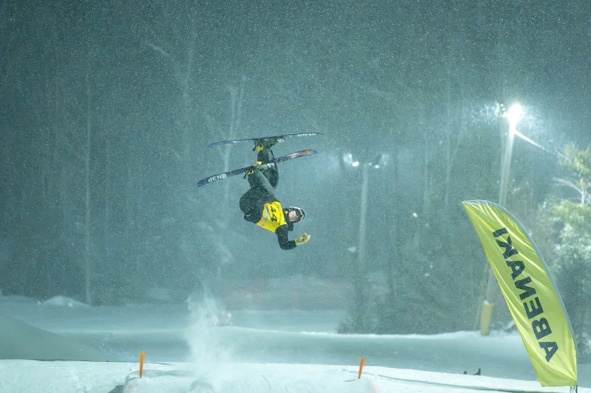 A freestyle skier upside down over the jump in falling snow at night, an Abenaki flag at the landing