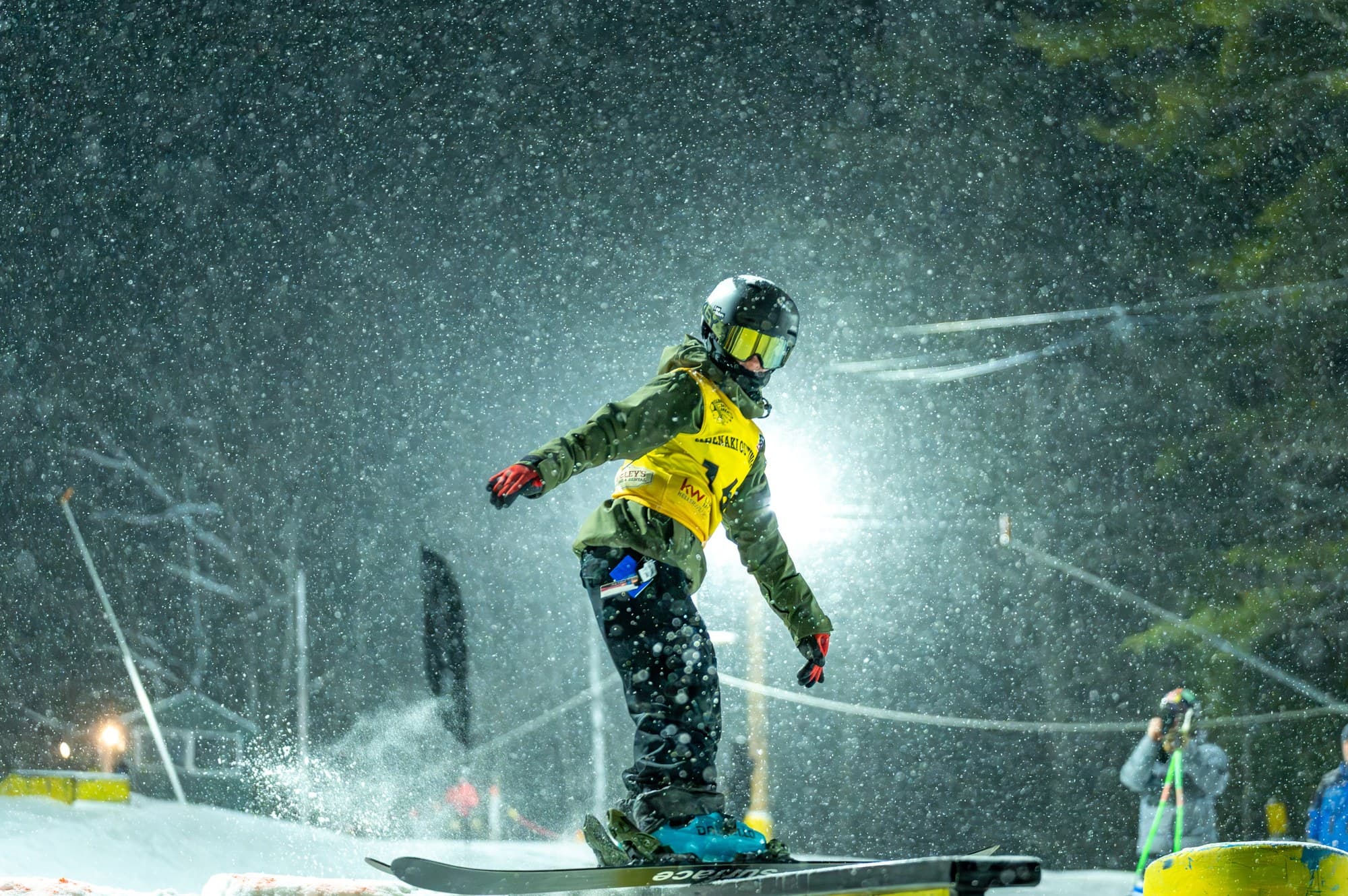 A young skier in a yellow bib riding a rail in the terrain park as snow falls under the lights