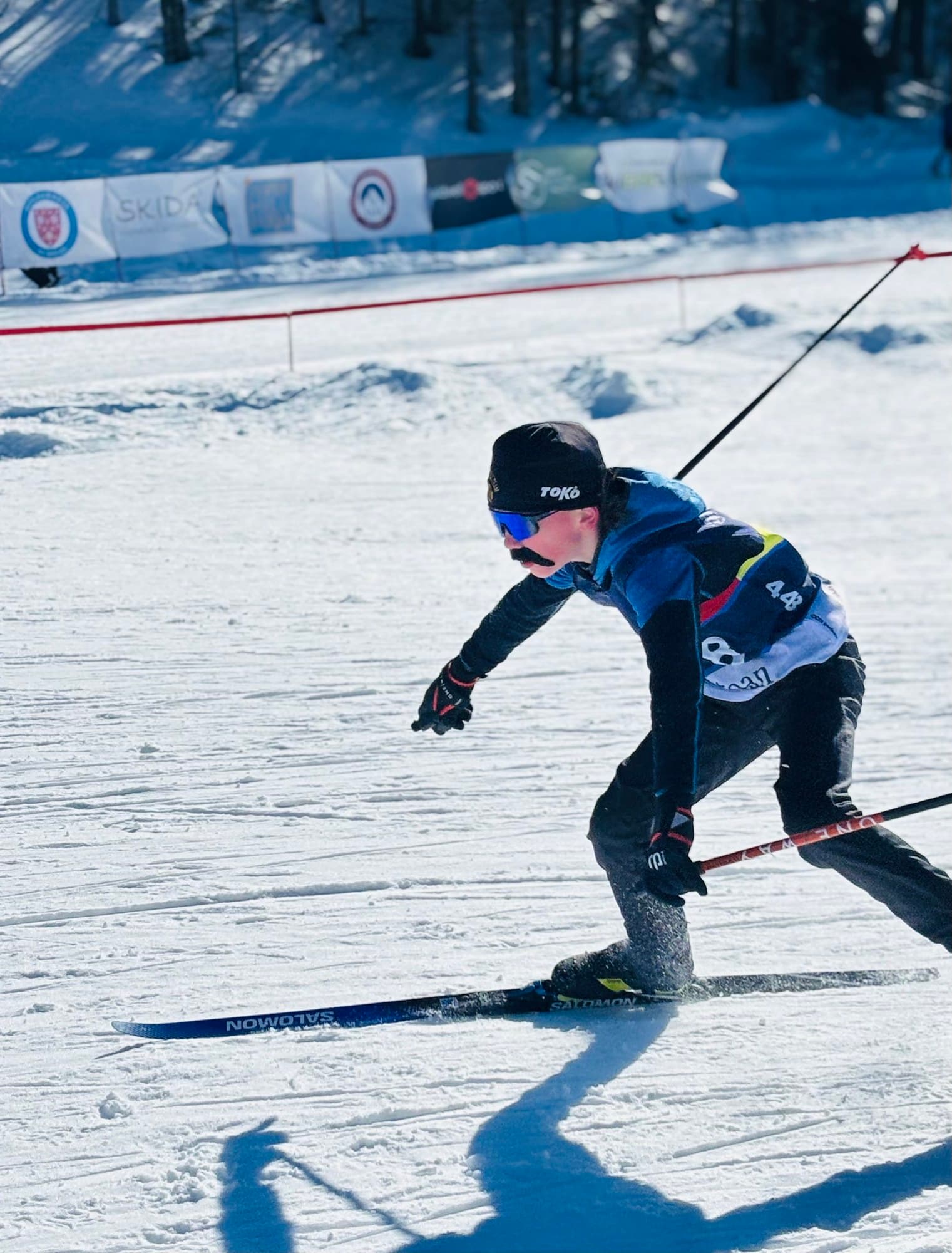 An Abenaki Nordic skier in a race bib skating hard past the sponsor banners at an away race