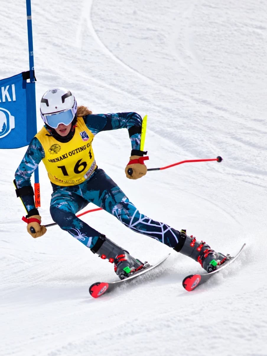 A U14 racer in Abenaki Outing Club bib 16 carving a giant slalom turn past a gate with the Abenaki goggle panel