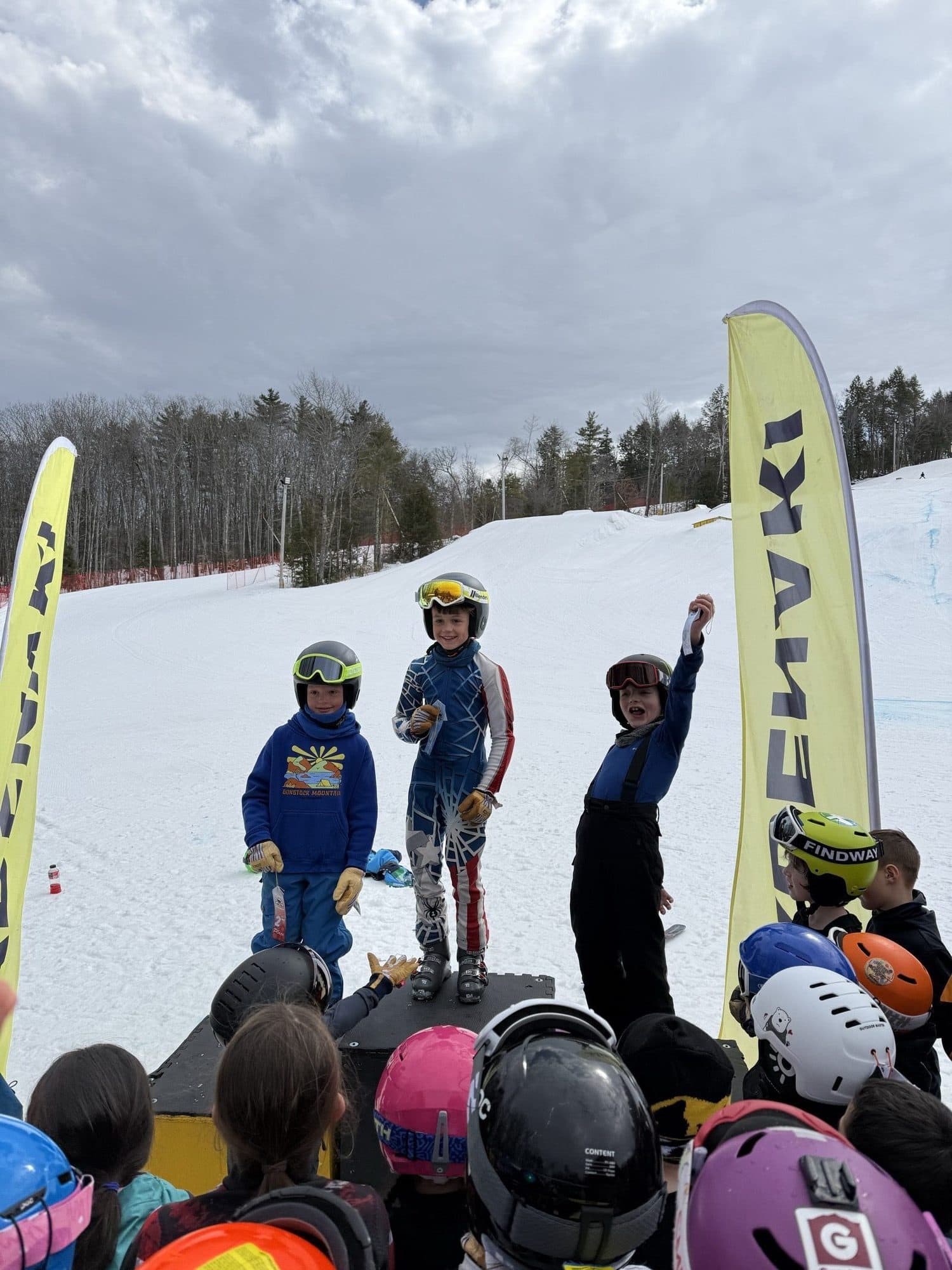 Three young athletes on the podium at a U8 race, watched by a crowd of teammates