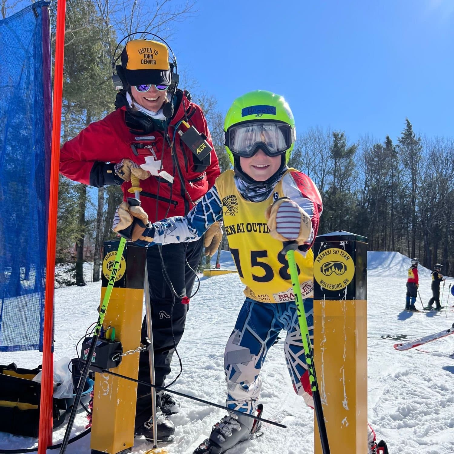 A U8 racer in a green helmet and Abenaki Outing Club bib 158 grinning in the start gate, the starter in a headset smiling behind