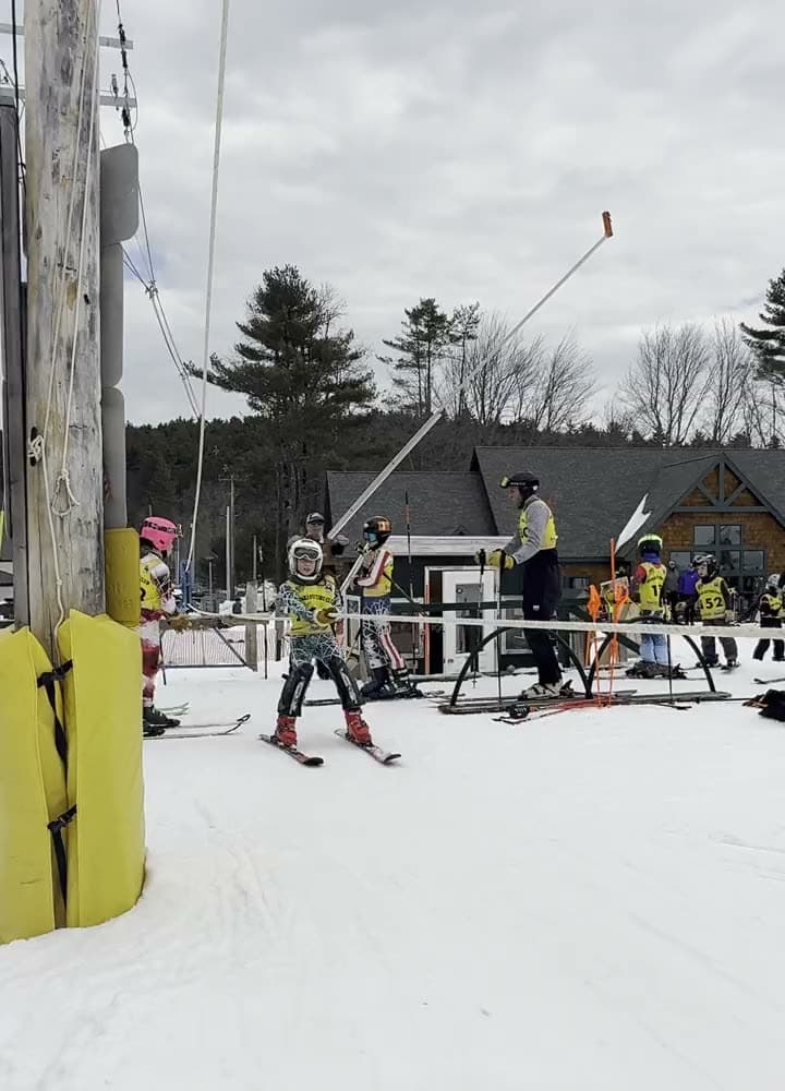 Young racers riding the rope tow up past the tower one after another, families gathered at the base of the hill, and a freestyle skier grabbing a ski high above the jump under the lights